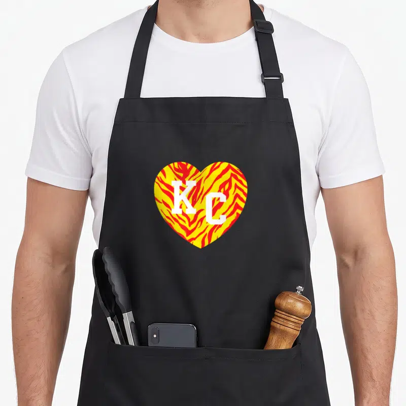 Kansas red striped apron - Heart shape with yellow and red tiger stripes containing white KC letters — shown on a black cotton canvas apron against a white background