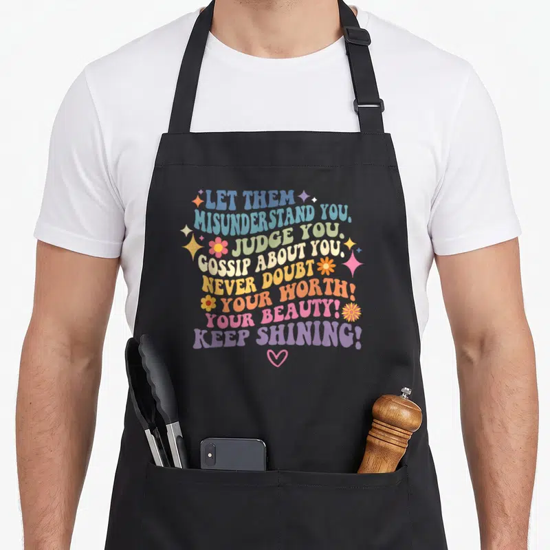 Them gossip apron - Stacked retro text in purple blue green yellow pink with small flowers stars and heart — shown on a black cotton canvas apron against a white background