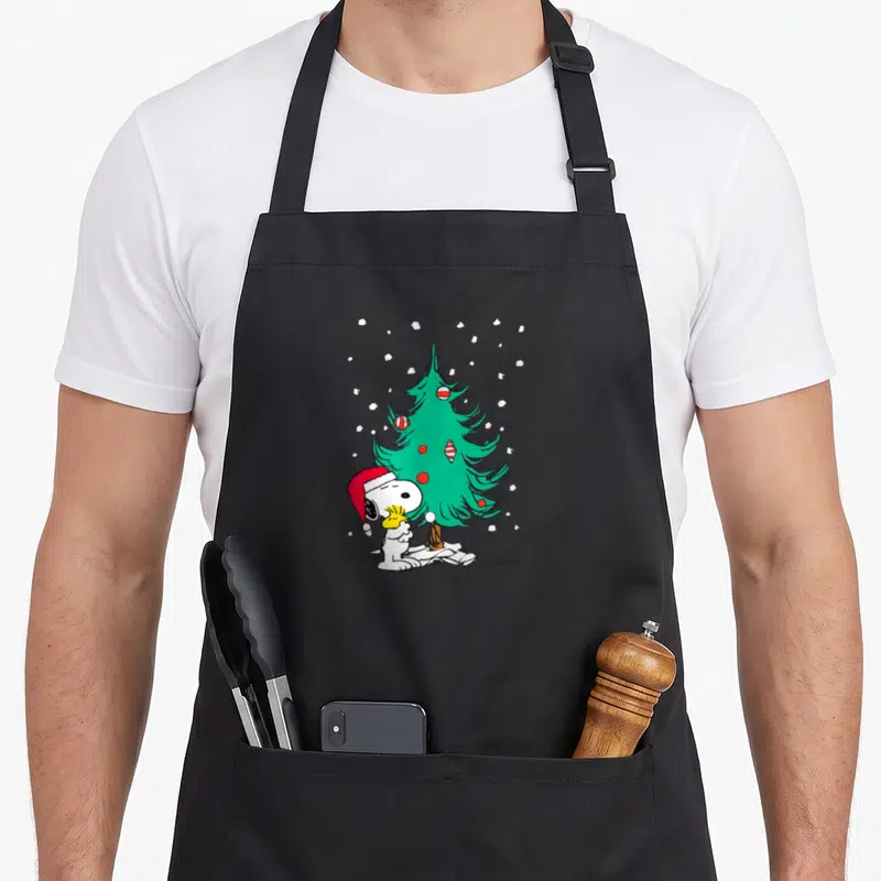 Cute holiday christmas tree apron - Snoopy in red hat sitting by green tree with red white ornaments and white — shown on a black cotton canvas apron against a white background