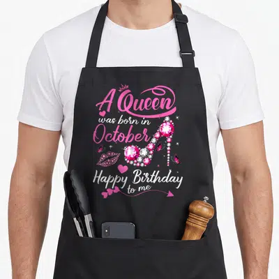 Funny october birthday apron - Pink white cartoon high heel shoe with A Queen was born in October text and jeweled — shown on a black cotton canvas apron against a white background