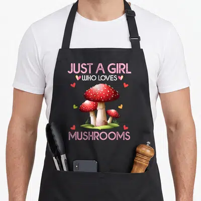 Vintage mushroom apron - Three red mushrooms with white spots surrounded by pink text and hearts — shown on a black cotton canvas apron against a white background