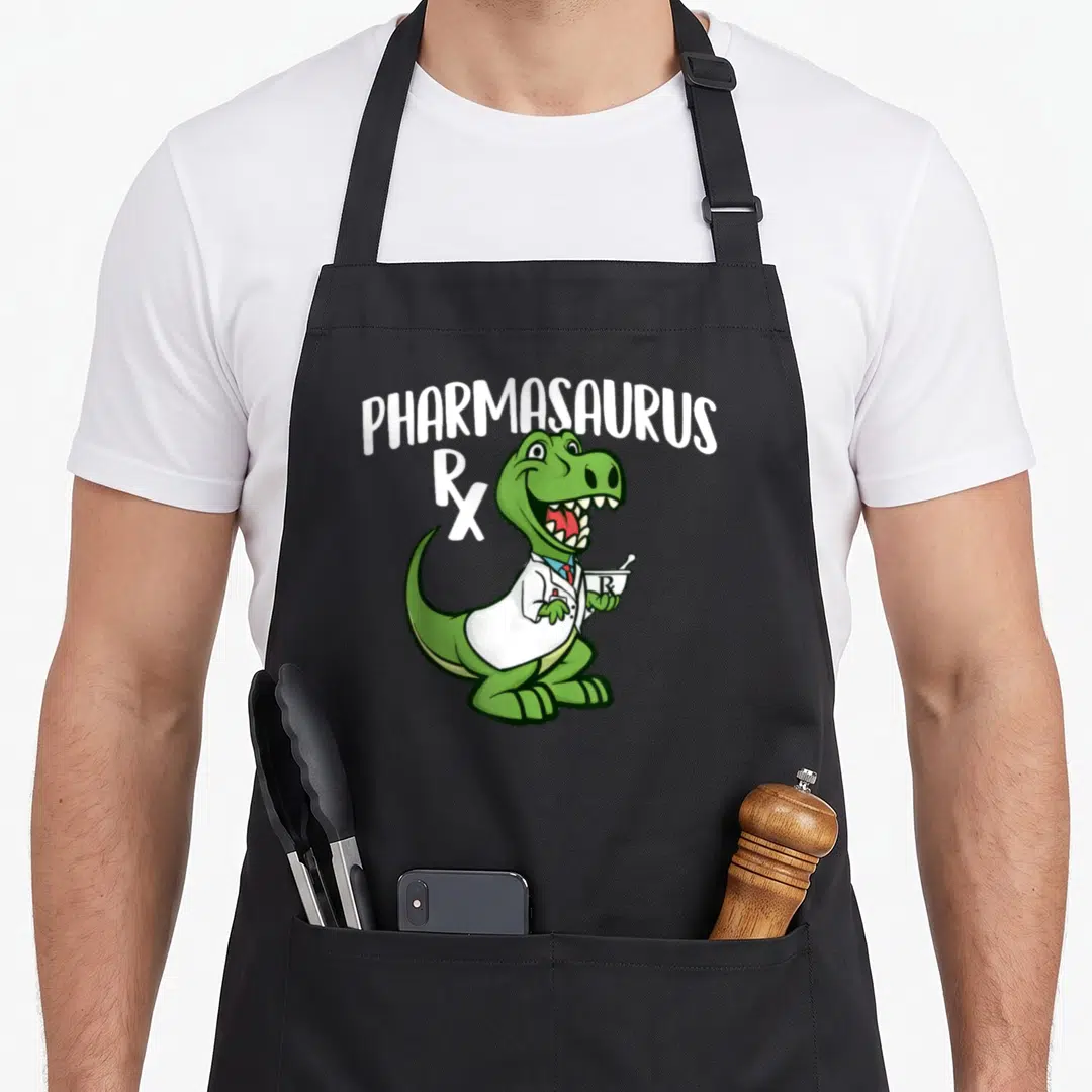 Pharmasaurus pharmacist apron - Green dinosaur in lab coat holding mortar pestle with PHARMASAURUS text and Rx symbol — shown on a black cotton canvas apron against a white background