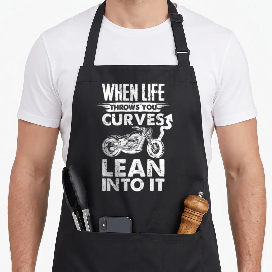 Throw motorcycle apron - White distressed text when life throws you curves lean into it above motorcycle with curving — shown on a black cotton canvas apron against a white background