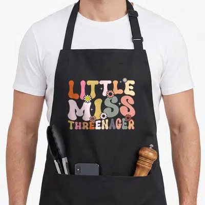 Miss boho apron - LITTLE MISS THREENAGER text with flowers stars and peace sign in orange pink yellow green blue purple — shown on a black cotton canvas apron against a white background