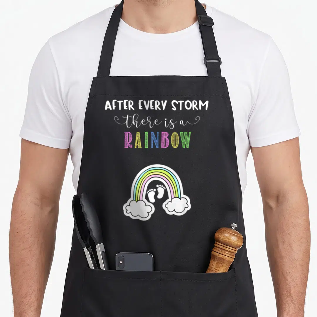 Pregnancy rainbow apron - Text reading After Every Storm There Is A Rainbow above rainbow arching over two clouds — shown on a black cotton canvas apron against a white background