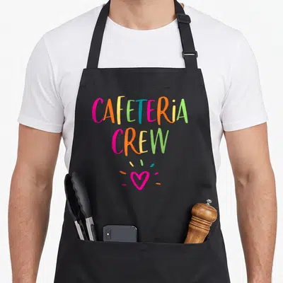 Appreciation cafeteria aprons - Stacked rainbow text reading CAFETERIA CREW above a pink heart with three scattered — shown on a black cotton canvas apron against a white background