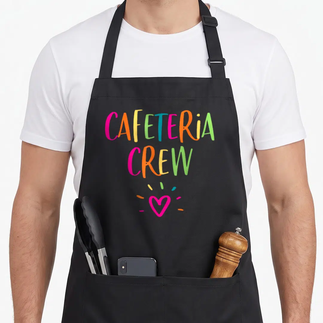 Appreciation cafeteria aprons - Stacked rainbow text reading CAFETERIA CREW above a pink heart with three scattered — shown on a black cotton canvas apron against a white background