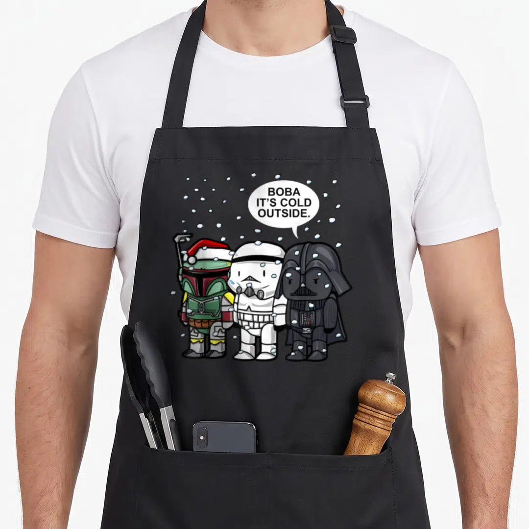 Boba christmas apron - Three chibi Star Wars characters under snowflakes with speech bubble saying Boba It's Cold — shown on a black cotton canvas apron against a white background