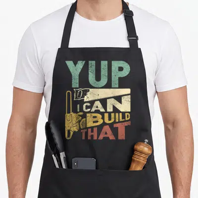 Build carpenter woodworking apron - Distressed YUP I CAN BUILD THAT text with yellow chainsaw and beige saw graphic — shown on a black cotton canvas apron against a white background