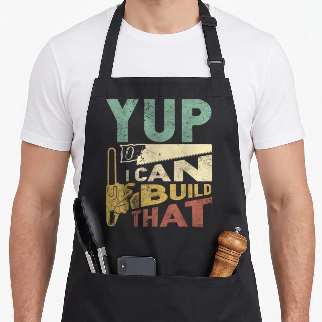 Build carpenter woodworking apron - Distressed YUP I CAN BUILD THAT text with yellow chainsaw and beige saw graphic — shown on a black cotton canvas apron against a white background