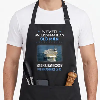 Independence father day apron - Aircraft carrier photo with white and blue text celebrating USS Independence CV-62 — shown on a black cotton canvas apron against a white background