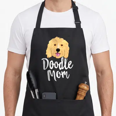 Doodle mother's apron - Cartoon dog head in tan and white above white Doodle Mom text — shown on a black cotton canvas apron against a white background