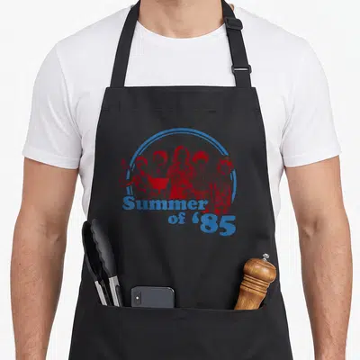 Shot summer aprons - Blue arc over five red figures above stacked Summer of '85 text — shown on a black cotton canvas apron against a white background