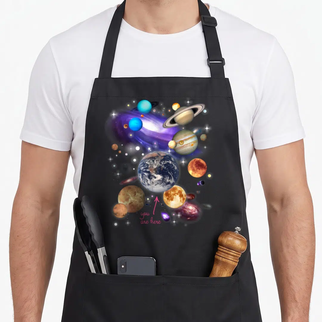 Solar system space planets apron - Earth surrounded by planets with pink arrow pointing to you are here text — shown on a black cotton canvas apron against a white background