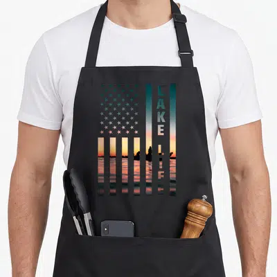 Lake american flag apron - USA flag shape with LAKE LIFE text and sunset boat silhouette in teal orange pink — shown on a black cotton canvas apron against a white background