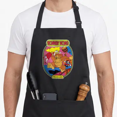 Cute donkey apron - Retro oval print with brown gorilla, blonde woman in red dress, man in blue and red, andDONKEY KONG — shown on a black cotton canvas apron against a white background