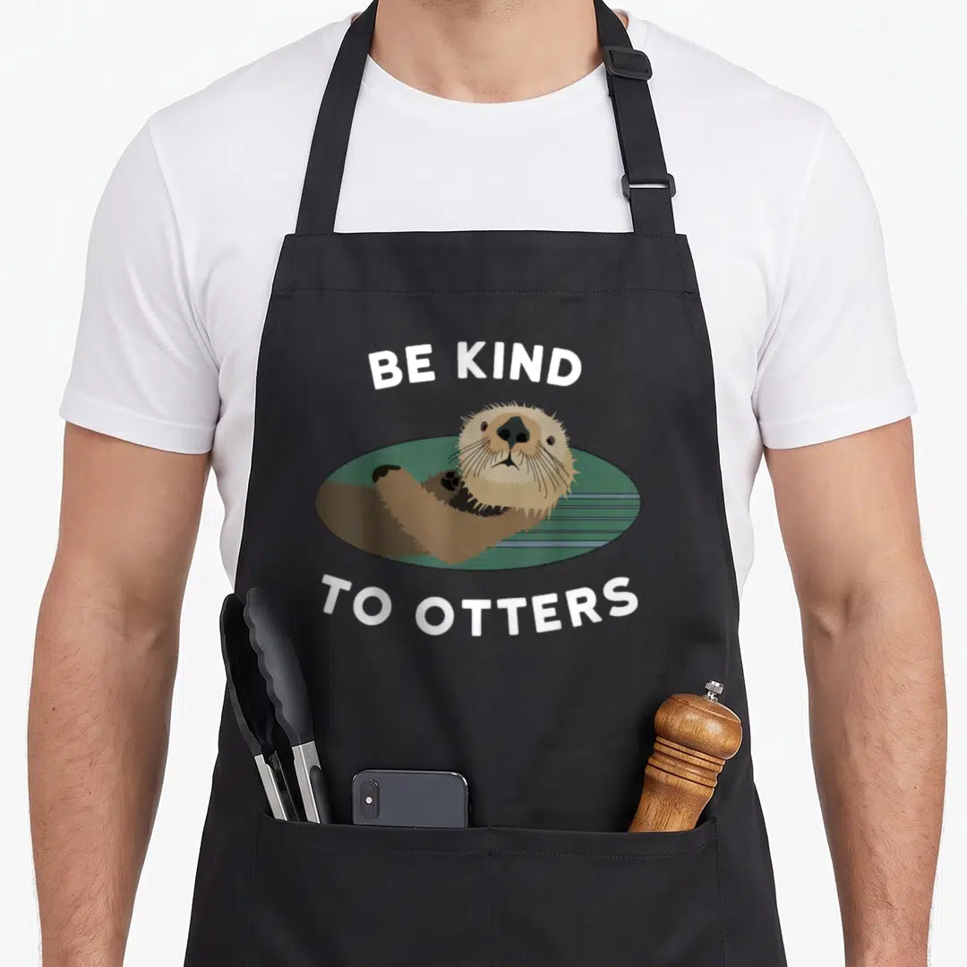 Otter pull girl apron - Cartoon brown otter in green water with white text BE KIND TO OTTERS — shown on a black cotton canvas apron against a white background