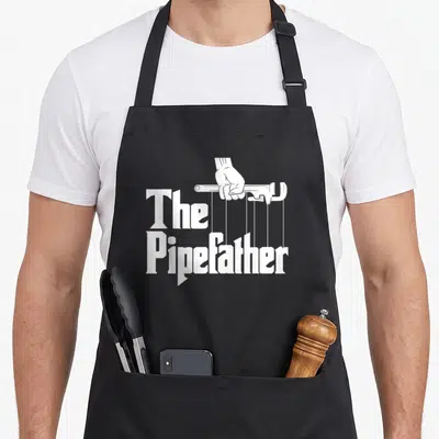 Plumbing pipefitter apron - White hand holding wrench with vertical lines connecting to The Pipefather text — shown on a black cotton canvas apron against a white background