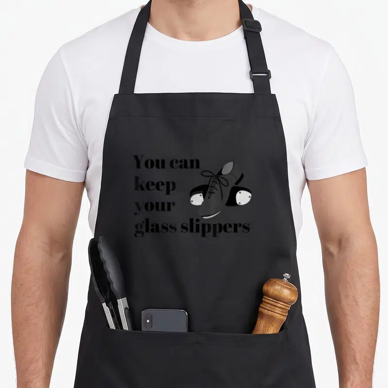 Tap dancing apron - Stacked text reading You can keep your glass slippers beside grey roller skate illustration — shown on a black cotton canvas apron against a white background