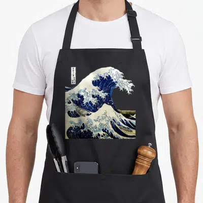 Wave japanese apron - Blue and white wave curling over boats with vertical text box and mountain background — shown on a black cotton canvas apron against a white background