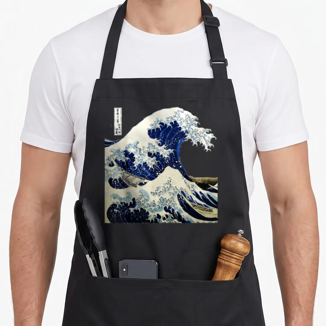 Wave japanese apron - Blue and white wave curling over boats with vertical text box and mountain background — shown on a black cotton canvas apron against a white background