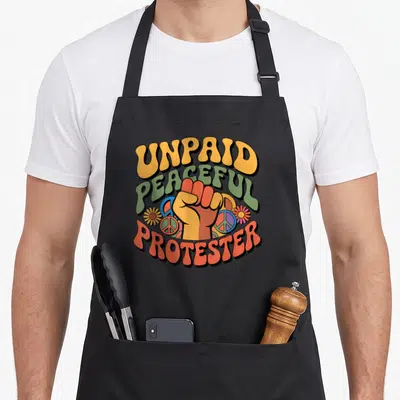Unpaid justice of the peace apron - Retro raised fist with UNPAID PEACEFUL PROTESTER text and peace symbols in yellow — shown on a black cotton canvas apron against a white background