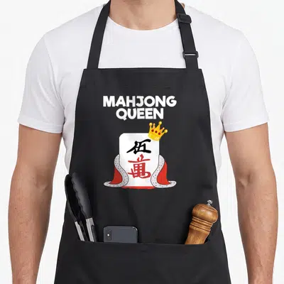 Queen mahjong apron - Stacked white text above crowned tile with red cape and yellow crown jewels — shown on a black cotton canvas apron against a white background