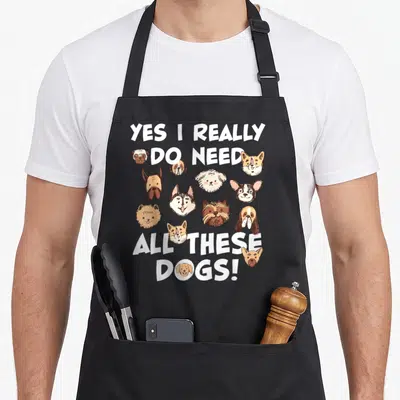 Rescue do i need an apron - YES I REALLY DO NEED ALL THESE DOGS text with cartoon dog heads in white brown and tan — shown on a black cotton canvas apron against a white background