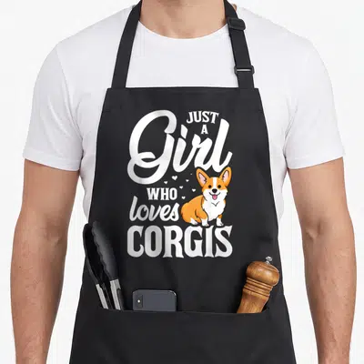 Corgis dog apron - Cartoon corgi with white text wrapping around it and small hearts scattered on grey background — shown on a black cotton canvas apron against a white background