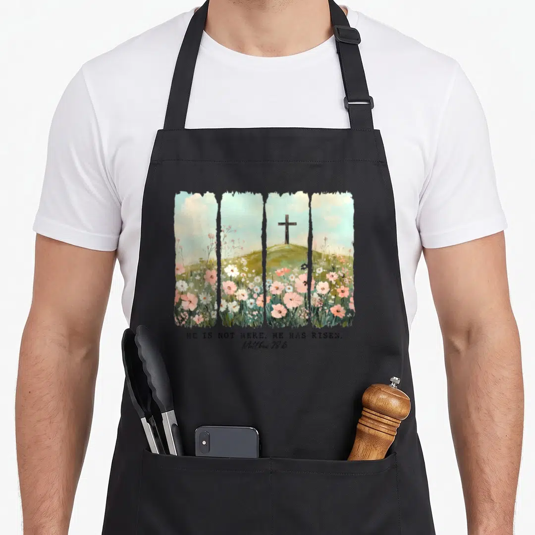 Christian he is risen apron - Watercolor cross on hill with pink and white wildflowers and Matthew 28:6 text — shown on a black cotton canvas apron against a white background