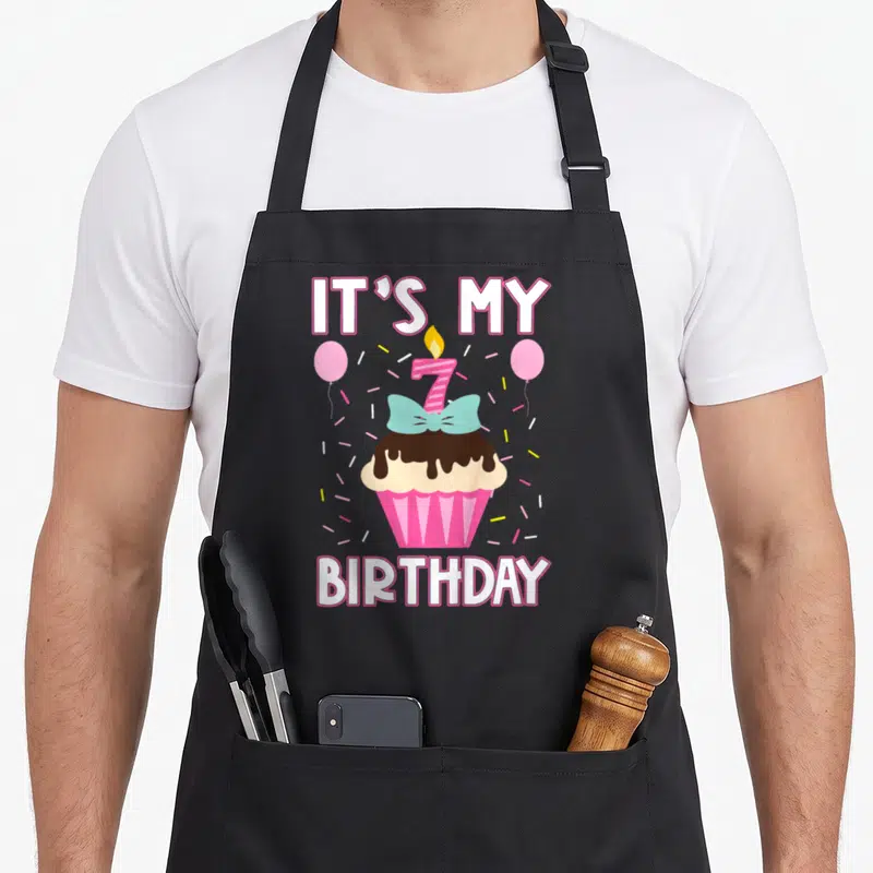 7th cupcake apron - IT'S MY 7 BIRTHDAY text above a pink cupcake with teal bow and lit candle surrounded by confetti — shown on a black cotton canvas apron against a white background