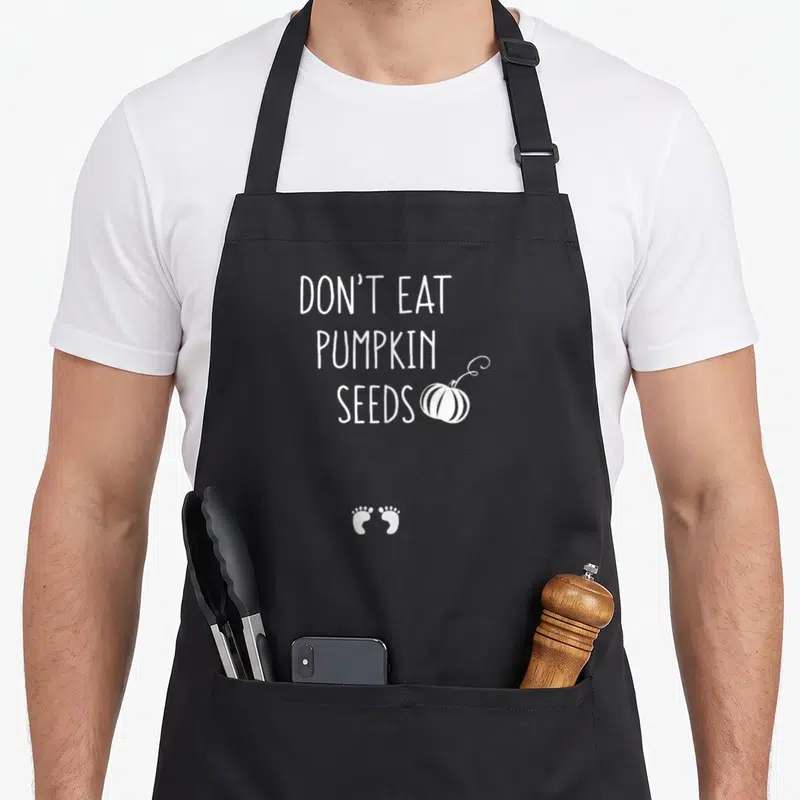 Eat thanksgiving apron - White text DON'T EAT PUMPKIN SEEDS with pumpkin and two baby footprints — shown on a black cotton canvas apron against a white background