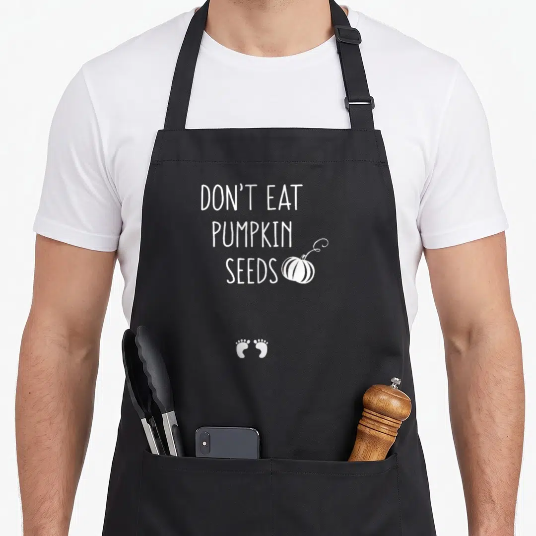 Eat thanksgiving apron - White text DON'T EAT PUMPKIN SEEDS with pumpkin and two baby footprints — shown on a black cotton canvas apron against a white background