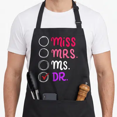 Doctorate doctor apron - Miss Mrs Ms Dr checklist with red checkmark in pink white and purple typography — shown on a black cotton canvas apron against a white background