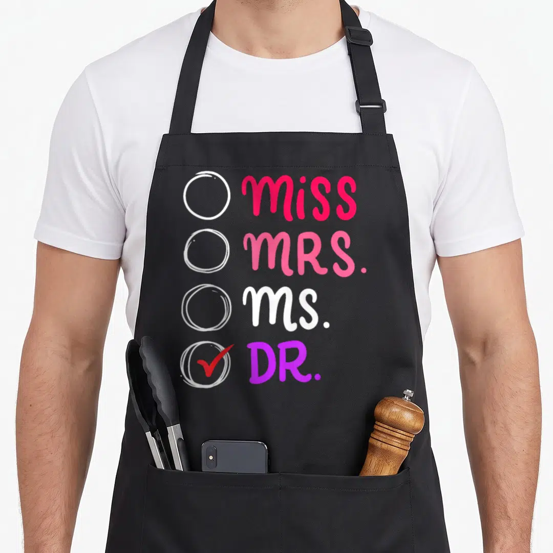 Doctorate doctor apron - Miss Mrs Ms Dr checklist with red checkmark in pink white and purple typography — shown on a black cotton canvas apron against a white background
