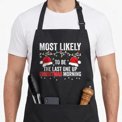 Most women's christmas apron - Stacked text reading Most Likely To Be The Last One Up with Santa hats and lights — shown on a black cotton canvas apron against a white background