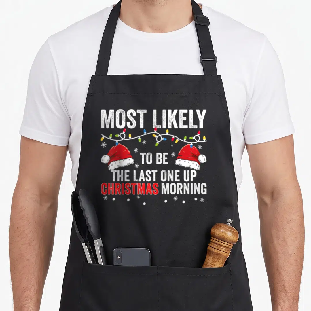 Most women's christmas apron - Stacked text reading Most Likely To Be The Last One Up with Santa hats and lights — shown on a black cotton canvas apron against a white background