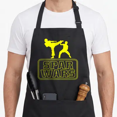 Funny taekwondo martial apron - Two yellow martial artist silhouettes above SPAR WARS text in rounded border — shown on a black cotton canvas apron against a white background
