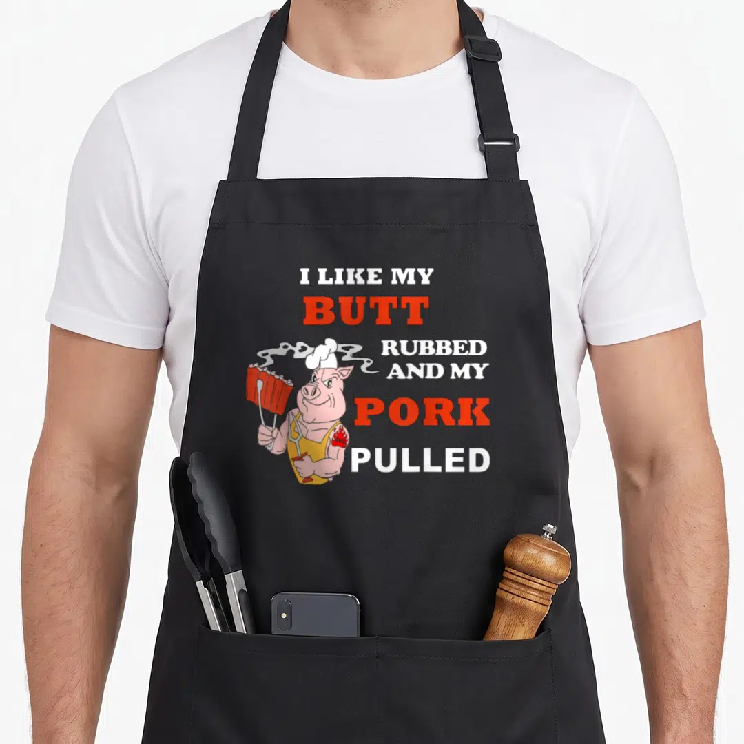 Butt summer mode apron - Cartoon pig chef holding ribs next to text I like my butt rubbed and my pork pulled — shown on a black cotton canvas apron against a white background