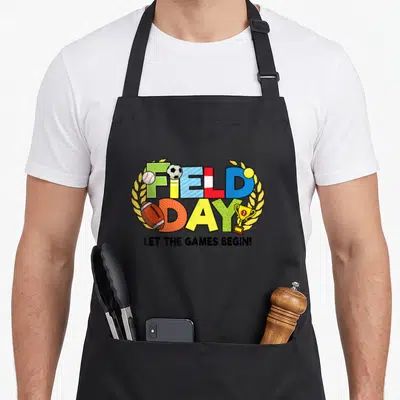 School field teacher apron - Colorful FIELD DAY text with baseball, soccer ball, tennis ball, football, and trophy — shown on a black cotton canvas apron against a white background
