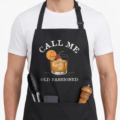 Recipe old fashioned aprons - Retro cocktail glass with CALL ME OLD FASHIONED text and orange slice garnish — shown on a black cotton canvas apron against a white background