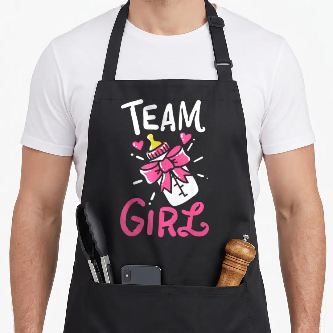 Birth gender reveal apron - Team Girl text above and below a pink-bowed baby bottle with two hearts — shown on a black cotton canvas apron against a white background
