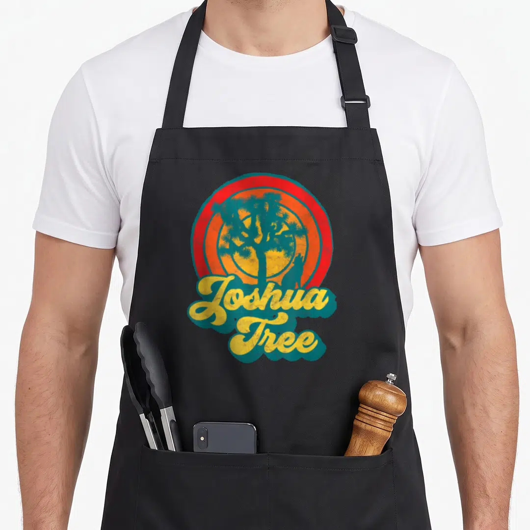 Coyote tree apron - Retro joshua tree and coyote silhouette against red orange concentric sun with yellow script text — shown on a black cotton canvas apron against a white background
