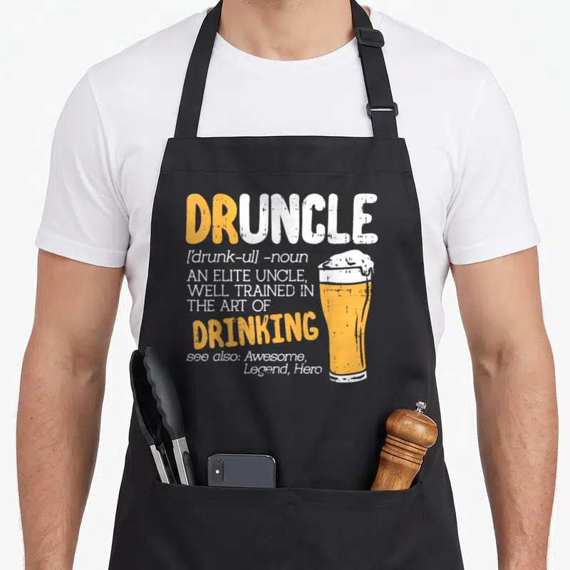 Boredkoalas art husband apron - Orange and white Druncle dictionary text next to a glass of beer with foamy head — shown on a black cotton canvas apron against a white background