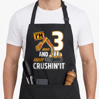 Excavator vehicle apron - Cartoon excavator overlapping number three with I'M 3 AND CRUSHIN' IT text — shown on a black cotton canvas apron against a white background