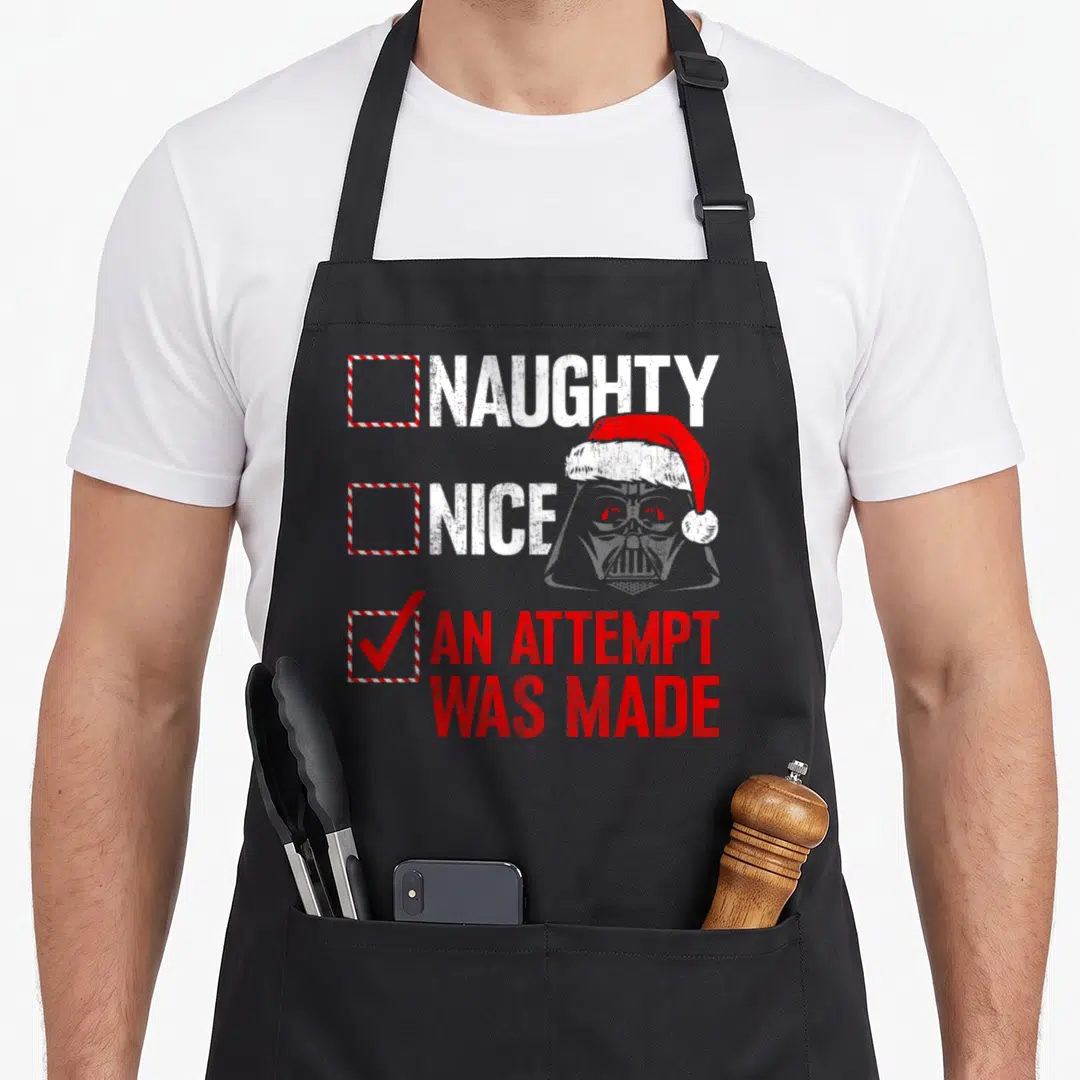 Naughty christmas apron - Darth Vader in Santa hat behind Naughty Nice An Attempt Was Made checklist with red checkmark — shown on a black cotton canvas apron against a white background