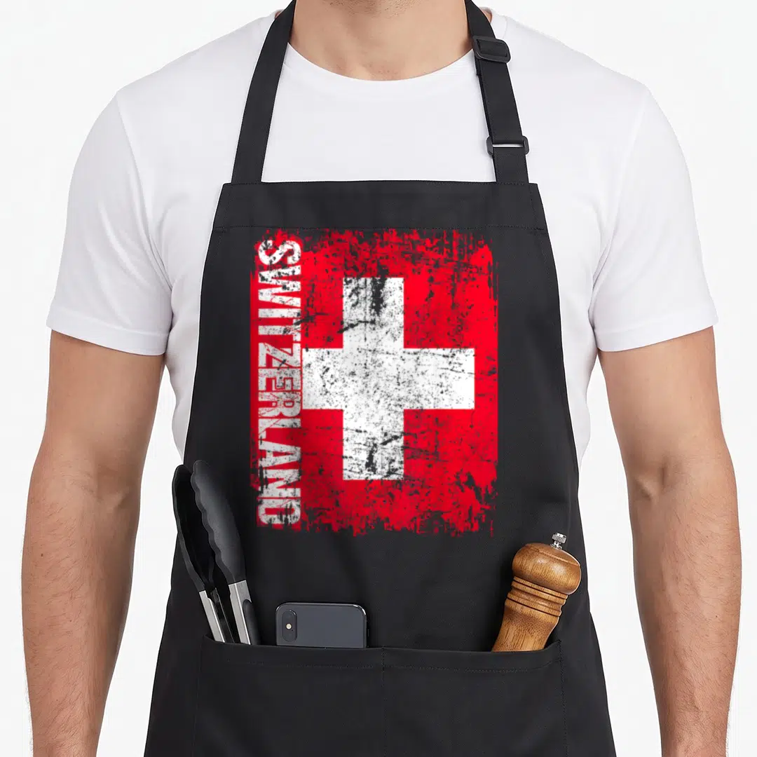 Distressed swiss apron - Red square with white cross and vertical SWITZERLAND text in distressed style — shown on a black cotton canvas apron against a white background