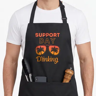 Support summer aprons - Retro sunglasses with palm tree reflections and SUPPORT DAY Drinking text in orange and grey — shown on a black cotton canvas apron against a white background