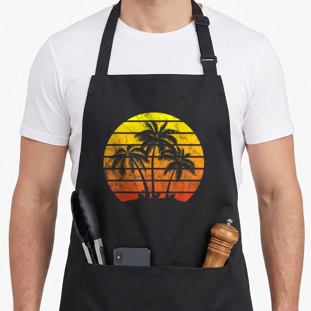 Sunset tree apron - Three palm tree silhouettes in front of a striped yellow, orange, and red retro sun — shown on a black cotton canvas apron against a white background