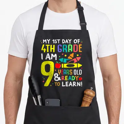 4th school grade apron - Stacked text with yellow pencil, red apple, rockets, hearts, stars in yellow red blue green — shown on a black cotton canvas apron against a white background